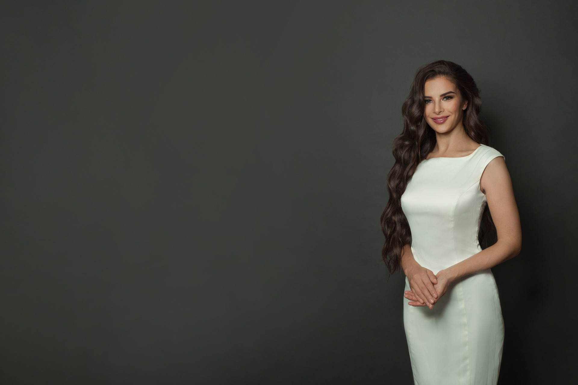 Happy smiling brunette woman with long shiny wavy hair in white silky dress posing on black studio background